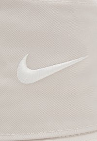 Light beige bucket hat made of soft fabric, featuring a white embroidered Nike logo on the front. Smooth texture with minimal stitching visible.