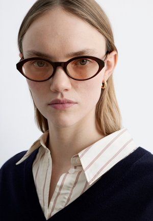 Jonge vrouw met lichtbruin haar die ovale, bruin getinte glazen draagt, een gouden creooloorbel, een marineblauwe trui en een wit shirt met dunne strepen.