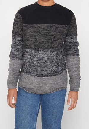Person trägt einen langärmeligen Strickpullover mit schwarzen, dunkelgrauen und hellgrauen horizontalen Farbstreifen, kombiniert mit blauen Jeans.