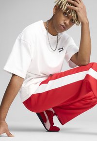 Hvid bomulds t-shirt med sort Puma-logo, parret med røde joggingbukser med hvide striber. Figuren har kort blond hår og en sølv kæde.
