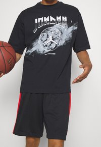 Black t-shirt with white "JORDAN" graphic, featuring a basketball design. Worn with black shorts having red side stripes. Holding a basketball.