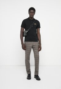 Fekete póló sárga logóval, kockás barna nadrág és fekete cipő. Kis fekete keresztpántos táska a vállon.