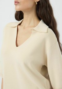 Beige neulepaita, jossa on V-pääntie, lyhyet hihat ja käännetty kaulus. Kangas on pehmeä ja tekstuuriltaan mielenkiintoinen. Minimalistiset suunnitteluyksityiskohdat.