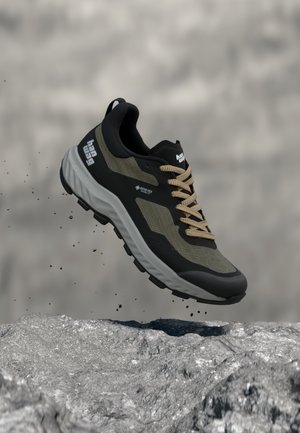 Črno-bežna Gore-Tex pohodniška obutev z grobo podplatom, v zraku nad skalnato površino z majhnimi kamni, ki padajo okoli nje.