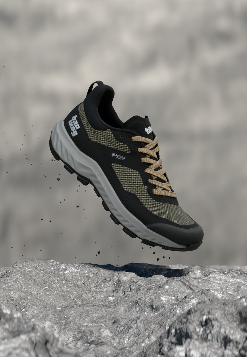 Μαύρο και μπεζ Gore-Tex πεζοπορικό παπούτσι με τραχιά σόλα, στον αέρα πάνω από βραχώδη επιφάνεια με πέτρες να πέφτουν γύρω του.