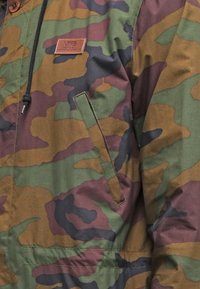 Bunda s maskárovým vzorem, s nakloněnou kapsou a koženým logem značky Vans na hrudi, vybavená černou šňůrkou s kovovým koncem.