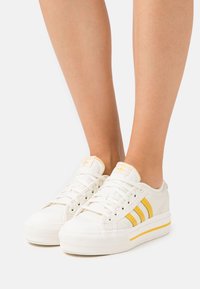 Bílé tenisky se třemi vertikálními žlutými pruhy, strukturovanou gumovou podešví a prošívanými detaily. Obsahují bílé tkaničky a značkový jazyk.