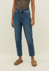 Persoon draagt een hoge taille blauwe jeans en bruine instap schoenen, staande tegen een effen lichte achtergrond.