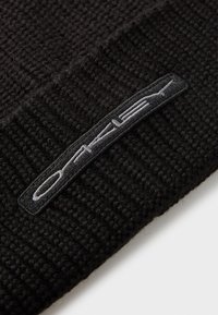 Black knit beanie with a textured weave. Features a prominent gray embroidered logo patch. Durable, flexible material. Simple design.