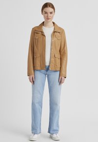 Hellbraune Lederjacke mit Kragendesign, zwei Fronttaschen und Druckknopfverschlüssen. Kombiniert mit hellblauen Jeans und weißen Turnschuhen.