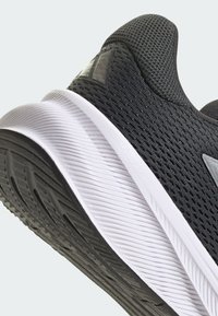 Chaussure de sport noire avec un dessus en mesh texturé, semelle intermédiaire blanche et semelle en caoutchouc. Présente un design minimaliste et un renfort au niveau du talon.