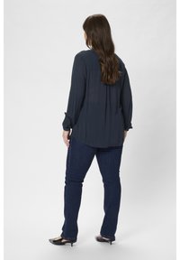 Blouse bleu foncé à manches longues avec un dos plissé, associée à un jean foncé. La blouse a une texture douce et des poignets boutonnés. Chaussures noires à talons.