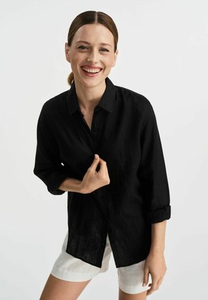 Czarna lniana koszula na guziki z podwiniętymi rękawami, w połączeniu z białymi szortami. Materiał wydaje się być teksturowany i lekki, odpowiedni na ciepłe dni.