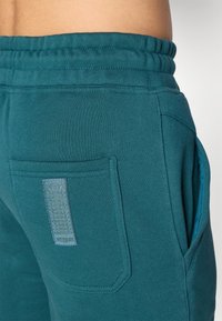 Calças de moletom verde-água com cintura elástica e bolsos laterais. Apresentam um detalhe texturizado nos bolsos em uma cor mais clara. Material suave, semelhante ao algodão.