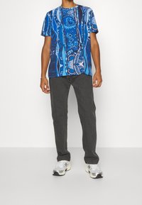 Man som bär en blå mönstrad kortärmad skjorta, mörkgrå jeans, vita sneakers och ett silverarmband, stående mot en vit bakgrund.