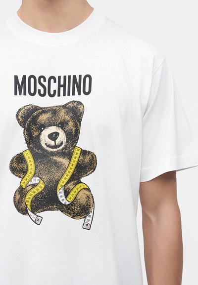 Λευκό μπλουζάκι με το κείμενο "MOSCHINO" πάνω από ένα καφέ αρκουδάκι τυλιγμένο με κίτρινη και λευκή μεζούρα.
