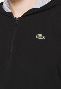 Černá mikina na zip s šedou podšívkou; na levé části hrudníku je malá vyšívaná zelená logo krokodýla. Měkký bavlněný materiál.