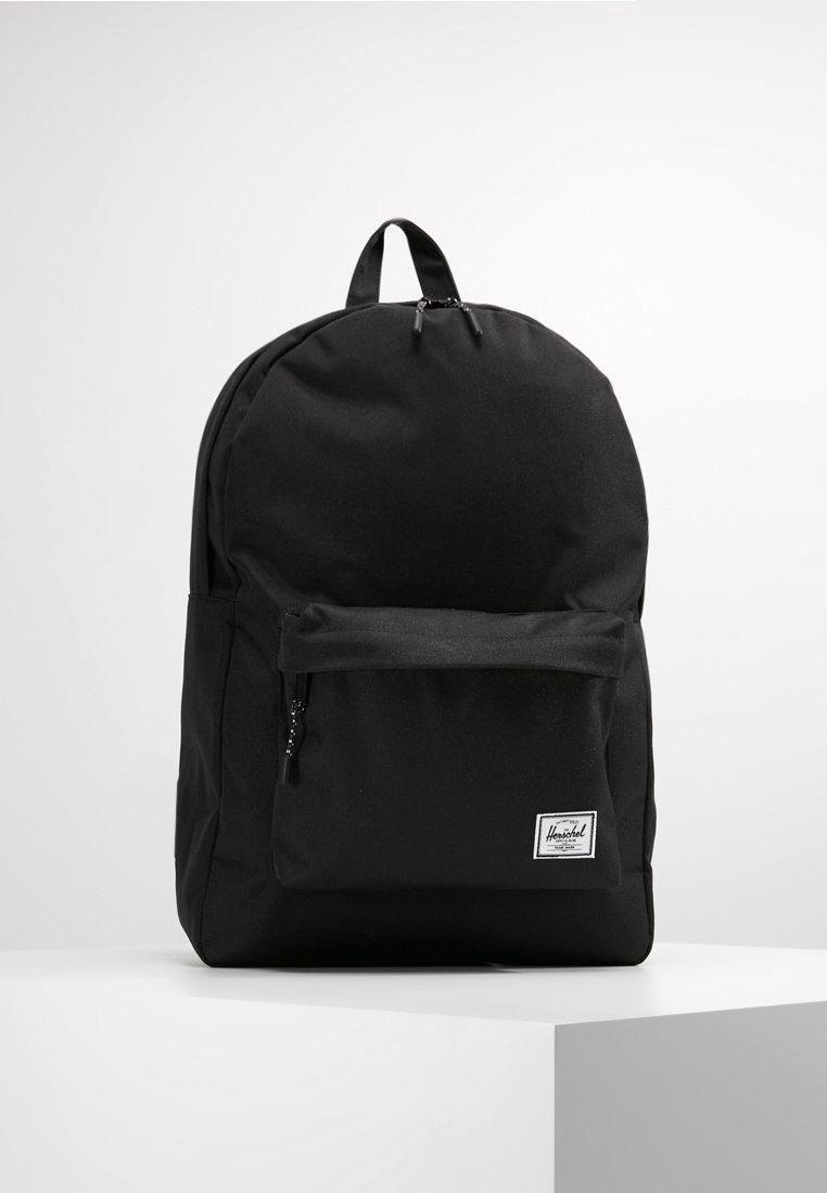 Sac à dos noir Herschel avec poche zippée à l'avant et poignée supérieure, présenté sur un socle blanc sur fond blanc uni.