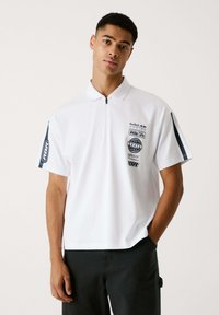 Camiseta polo blanca de manga corta, con cuello con cremallera y logotipos de Red Bull Racing en el pecho y las mangas. Tejido suave con un ajuste relajado.