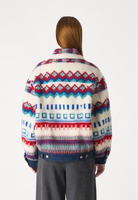 Chaqueta de forro polar con base blanca y patrones geométricos coloridos en rojo, azul y verde azulado, y un bajo de mezclilla con botones metálicos.