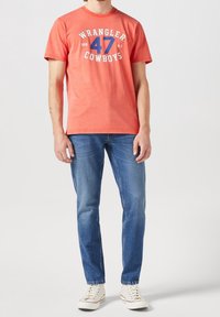 Röd bomullst-shirt med "WRANGLER 47 COWBOYS"-logotyp i blått och vitt; bärs med blå denimjeans och vita sneakers.