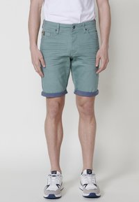 Shorts vert menthe avec une finition texturée, ourlet roulé, quatre poches et une fermeture à bouton à l'avant. Associés à des chaussures de sport blanches.
