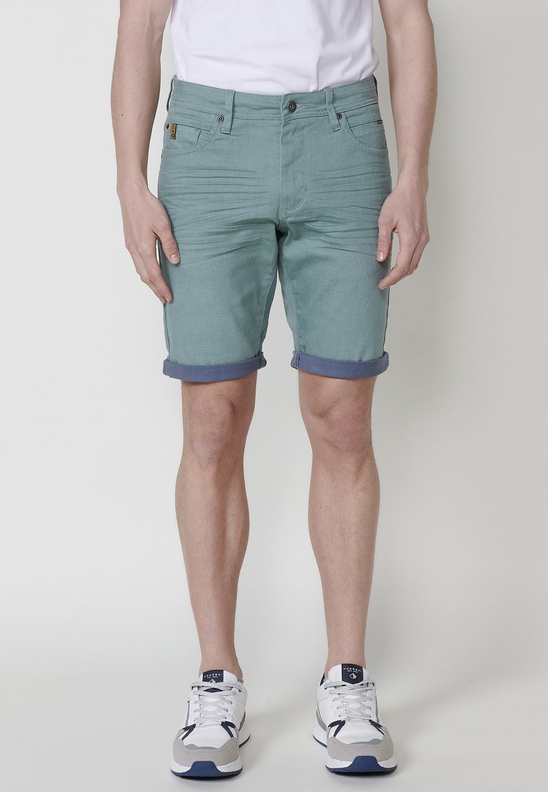 Shorts vert menthe avec une finition texturée, ourlet roulé, quatre poches et une fermeture à bouton à l'avant. Associés à des chaussures de sport blanches.