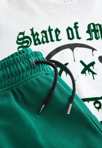 Pantalon de jogging en coton vert avec une taille élastique et un cordon de serrage noir, associé à un T-shirt blanc arborant un texte graphique vert et des motifs.