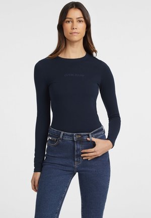 Navy long-sleeve bodysuit with a fitted design, soft material, and subtle "GUESS JEANS" branding on the front, paired with high-waisted denim jeans.