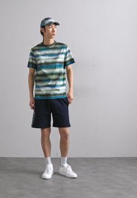 Μπλουζάκι κοντό μανίκι με σχέδιο από πράσινα και μπλε κύματα, συνδυασμένο με ναυτικές σορτς και άσπρα αθλητικά παπούτσια. Το καπέλο ταιριάζει με τα χρώματα της μπλούζας.