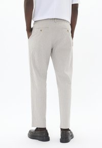 Pantalones de rayas de color claro con un ajuste relajado, que cuentan con bolsillos laterales y traseros, y sutiles detalles de botones en tono dorado. Llevados con zapatos oscuros.