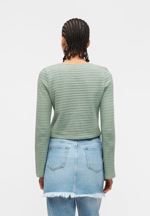 Personne avec des cheveux tressés portant un haut vert texturé à manches longues et une jupe en jean bleu clair effilochée par-dessus un jean, vue de dos.