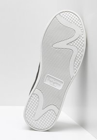 Czarny but z białą gumową podeszwą, z widocznymi teksturowanymi wzorami i wytłoczonym logo. Posiada falisty kształt z okrągłymi uchwytami dla lepszej przyczepności.