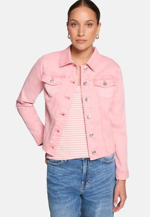 Femme portant une veste en jean rose clair boutonnée par-dessus une chemise rayée rose et blanche avec un jean bleu sur fond uni.