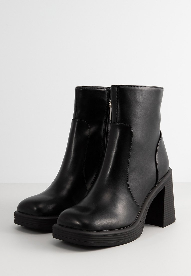 Une paire de bottines en cuir noir avec des talons hauts épais et des zips sur les côtés, sur une surface blanche.