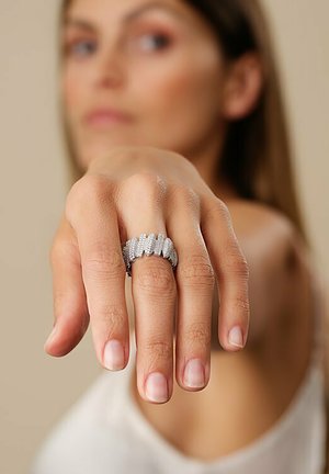 Silberfarbener Ring mit wellenförmigem Design, der mehrere kleine funkelnde Steine in einem strukturierten Band umfasst und an einem Finger getragen wird.