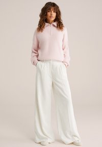 Gaiši rozā, teksturēts džemperis ar rāvējslēdzēja apkakli; kombinēts ar platām, baltām, ribotām biksēm. Modelis stāv ar rokām kabatās pret neitrālu fona.