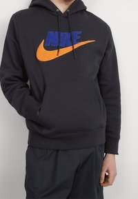 Sudadera negra con un gran logo azul de "NIKE" y un swoosh naranja. Presenta un bolsillo frontal y capucha con cordones. Tejido de textura suave.