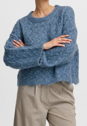 Personne portant un pull bleu à torsades avec les bras croisés et un pantalon beige à rayures fines, debout devant un fond uni.