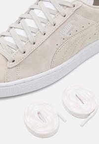 Világos bézs suéd sneaker fehér talppal, textúrált részletekkel és fehér, pasztell csíkkal díszített lapos fűzőkkel. Két további, hozzáillő fűző is tartozik hozzá.