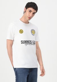 Tricou alb din bumbac, cu mâneci scurte, având logo-uri circulare galbene și negre și textul negru "SUNNIES" pe față. Purtați cu blugi albaștri.