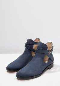 Un par de botines de ante azul marino con suelas negras y correas con hebilla envolventes, mostrados sobre una superficie blanca contra un fondo gris claro.