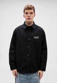 Μαύρο πουκάμισο με κουμπιά κατασ�κευασμένο από λείο ύφασμα, με κεντημένο λογότυπο στην αριστερή πλευρά του στήθους και κλασικό γιακά.