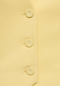 Žlutý blejzr s hladkým texture, který má tři kulaté látkové knoflíky po levé straně, prošité knoflíkové dírky a přizpůsobený tvar.