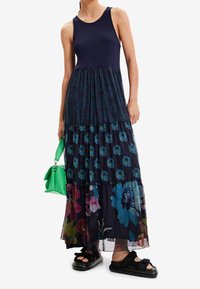 Robe maxi avec un haut côtelé bleu marine et une jupe florale superposée aux teintes sombres, présentant des motifs bleus et roses. Accompagnée de sandales noires et d'un sac à main vert.