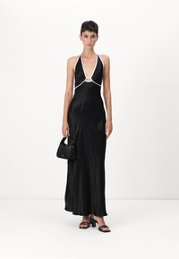 Black satin maxi dress with halter neck, white accents, and circular cutout. Paired with a small black handbag and strappy block-heeled sandals.