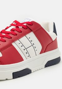 Baskets rouges et blanches avec une tige en cuir lisse, présentant un design rayé et un logo "TOMMY JEANS". Semelle intermédiaire blanche, accent bleu marine sur la semelle extérieure.