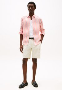 Chemise à rayures rouges et blanches à manches longues, débardeur blanc, shorts beiges et mocassins noirs. Tenue décontractée avec une coupe ample et des boutons.