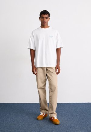 Giovane uomo in piedi su un tappeto blu che indossa una maglietta bianca oversize, pantaloni beige e sneaker arancioni davanti a una parete bianca.