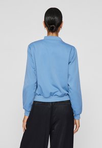 Modra, lahka jakna s ovratnikom in elastičnim robom, dolge rokave in gladko teksturo. Obučena z črnimi hlačami.